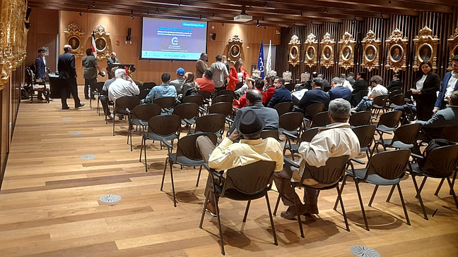 A formal event in a large auditorium filled with people seated in chairs. A presentation is visible on a screen at the front of the room. There’s a person standing and speaking.