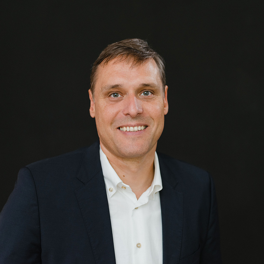 Ein Mann mit kurzem, braunem Haar lächelt freundlich in die Kamera. Er trägt ein weißes Hemd und einen dunkelblauen Anzug. Der Hintergrund ist schlicht und schwarz, was den Fokus auf die Person lenkt. 