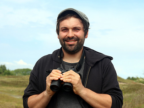 Franz Löffler stands outdoors, holding binoculars and smiling at the camera.