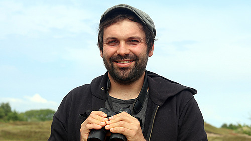 Franz L&ouml;ffler steht im Freiland, h&auml;lt ein Fernglas und l&auml;chelt in die Kamera