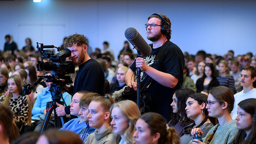 A cameraman and a sound engineer are standing in a hall between rows of spectators.