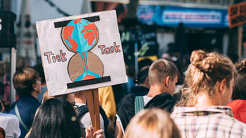 Menschen sind von hinten in einem Demonstrationszug zu sehen, zentral zu sehen ist ein Schild mit der Weltkugel als auslaufende Sanduhr.