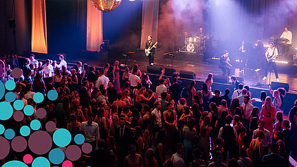 View from the gallery of a band on stage and the packed dance floor. A huge chandelier hangs from the ceiling. In two corners of the picture are colourful confetti circles as a frame.