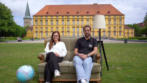 Ein Mann und eine Frau sitzen auf einem Sofa auf einer Wiese, im Hintergrund ist ein Schloss