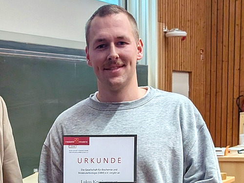A man with short blond hair smiles at the camera and holds a certificate in his hand.