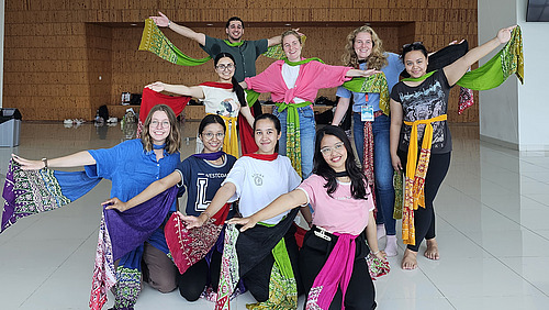 Eine Gruppe von jubelnden Frauen posiert fr&ouml;hlich f&uuml;r ein Foto, einige mit erhobenen Armen. Die lockere Pose und die bunten Kleider vermitteln eine fr&ouml;hliche und energiegeladene Atmosph&auml;re.
