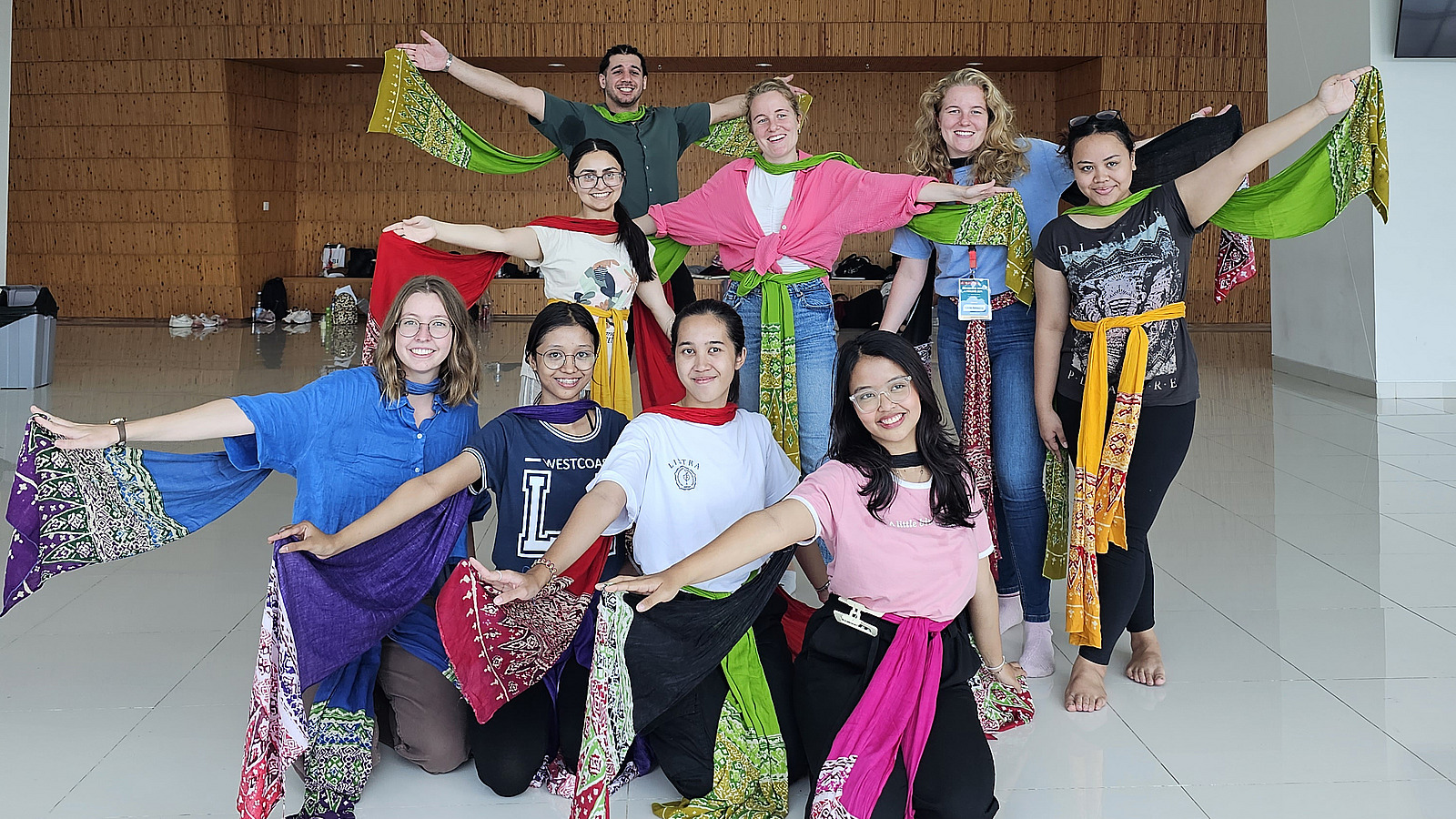 Eine Gruppe von jubelnden Frauen posiert fr&ouml;hlich f&uuml;r ein Foto, einige mit erhobenen Armen. Die lockere Pose und die bunten Kleider vermitteln eine fr&ouml;hliche und energiegeladene Atmosph&auml;re.