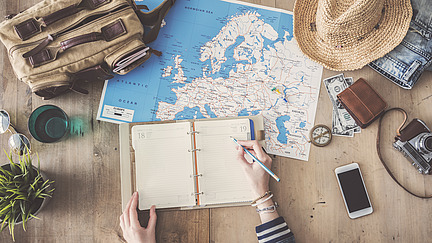 Symboldbild für die Vorbereitung einer Reise. Hut, Mobilphone, Kamera, Geldbeutel und eine Tasche liegen neben einer Landkarte von Europa und eine Person macht Notizen in einem kleinen Terminkalender.