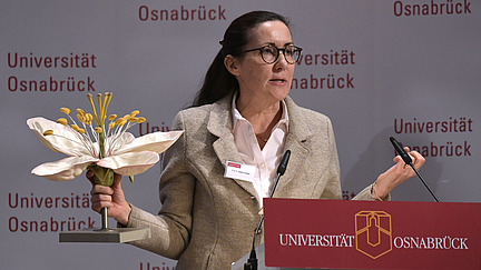 A woman is standing at a lectern, holding a large model of a flower in her hand.