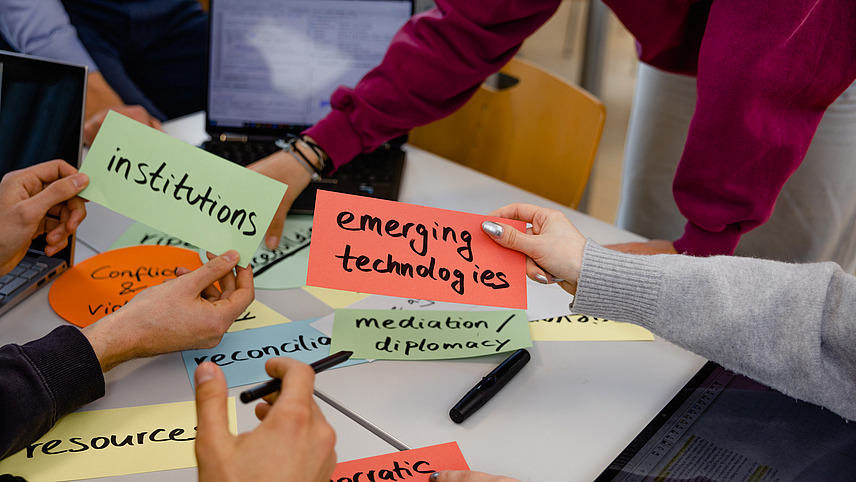 Viele H&auml;nde &uuml;ber einem Tisch tauschen bunte Karten mit Stichworten aus wie "emerging technologies", "democratic backsliding" oder "institutions"