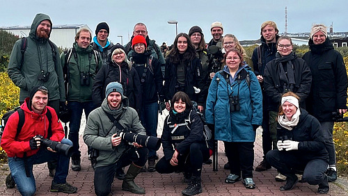A group of people stand outside wearing jackets and smiling. Some are carrying cameras and binoculars.