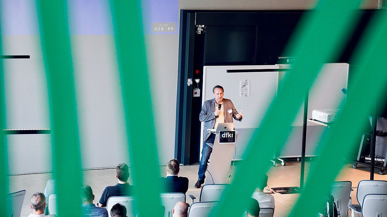 The picture shows a lecture hall in which a man is standing at a lectern and speaking in front of an audience. The view of the scene is framed by green vertical lines, which creates a special perspective.