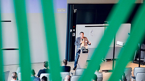 Das Bild zeigt einen Vortragssaal, in dem ein Mann an einem Rednerpult steht und vor Publikum spricht. Der Blick auf die Szene ist durch grüne vertikale Linien gerahmt, was eine besondere Perspektive erzeugt.