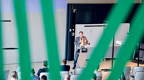 The picture shows a lecture hall in which a man is standing at a lectern and speaking in front of an audience. The view of the scene is framed by green vertical lines, which creates a special perspective.