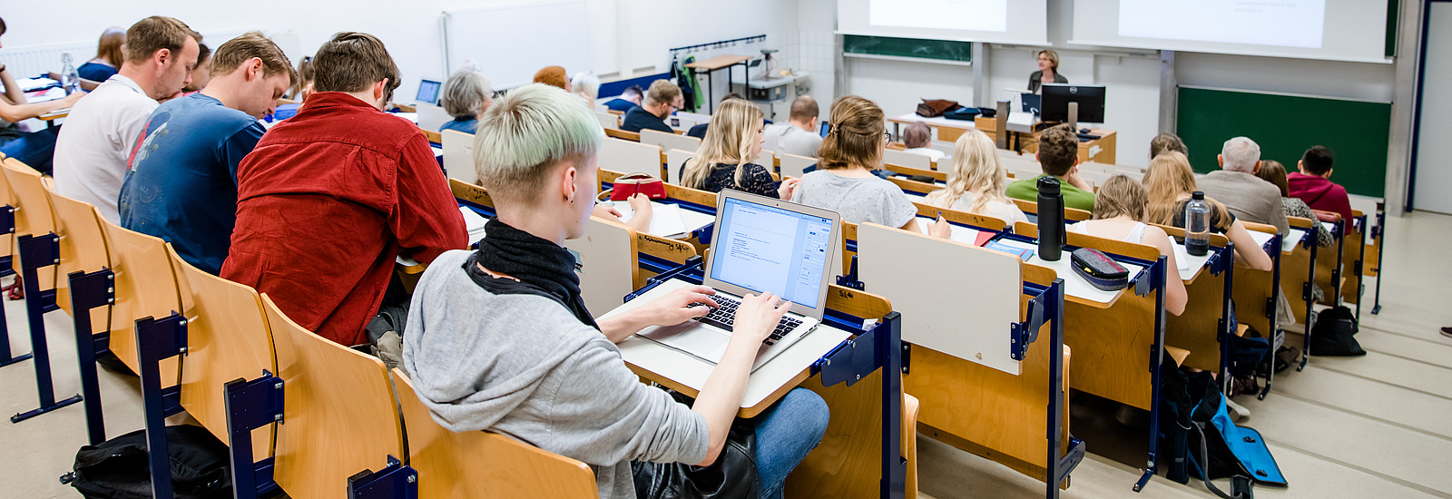 Blick von der Seite &uuml;ber die Sitzreihen eines kleinen H&ouml;rsaals auf Studierende. Vorne wird eine Pr&auml;sentation auf zwei W&auml;nde projeziert.