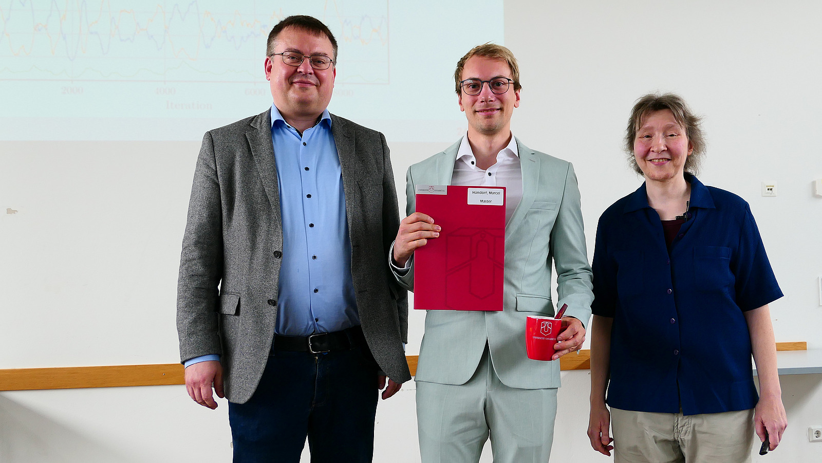 Ein Student mit Studiendekanin und Dekan stehen nebeneinander. Der Student hat eine Mappe in der Hand