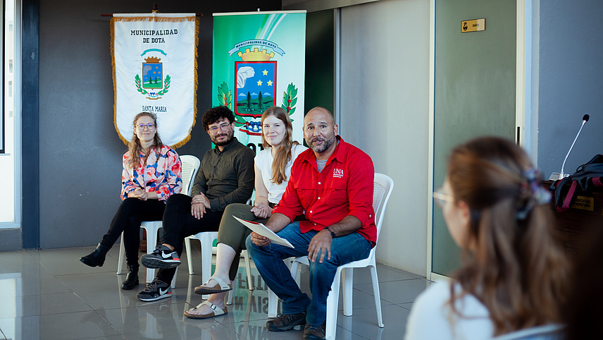 A panel discussion in a room. Four people are sitting on chairs, with two banners displaying symbols and text visible in the background. The person on the far right is holding a sheet of paper. Some members of the audience are visible in the foreground. 
