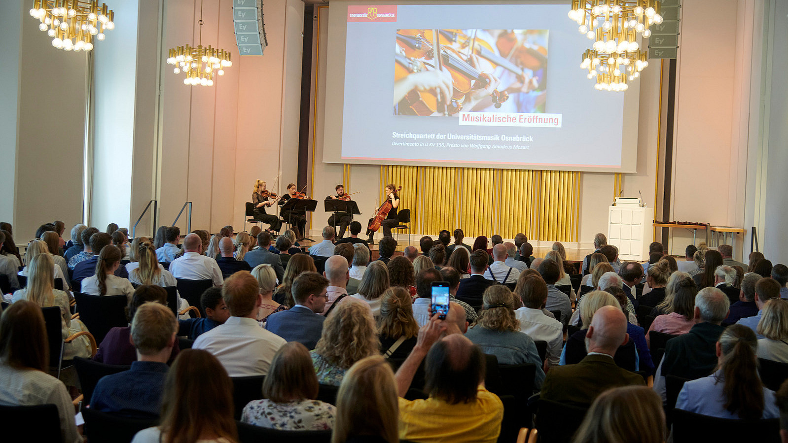 Four people with string instruments on a stage. Numerous people are sitting in the audience.