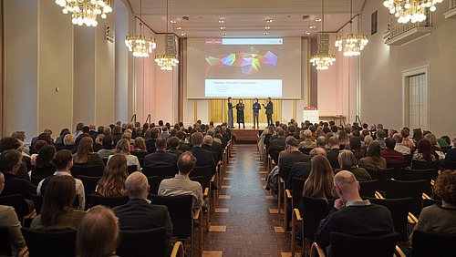 A choir is singing on stage in a large hall. Many people are sitting in the audience.