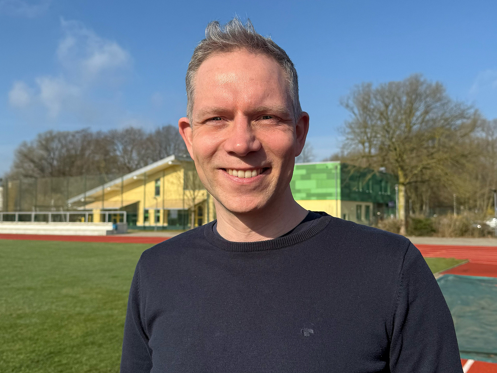Ein l&auml;chelnder Mann vor dem Hintergund eines Sportplatzes.