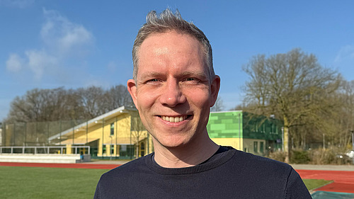 Ein l&auml;chelnder Mann vor dem Hintergund eines Sportplatzes.