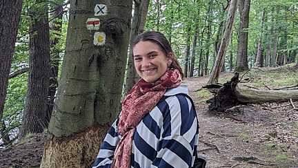 Eine junge Frau mit zusammengebundenem, dunklem Haar lächelt strahlend in die Kamera. Sie trägt einen gestreiften Mantel und einen roten Schal. Im Hintergrund ist ein bewaldeter Weg zu sehen, und neben ihr steht ein Baum mit aufgemalten Wandermarkierungen.