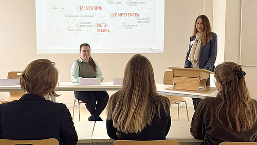 Blick in einen Veranstaltungsraum. Drei Personen sitzen an Seminarstischen und schauen auf eine Präsentation. Eine Person steht an einem Rednerpult, eine weitere sitzt vorne, und schaut die Anderen lächelnd an.