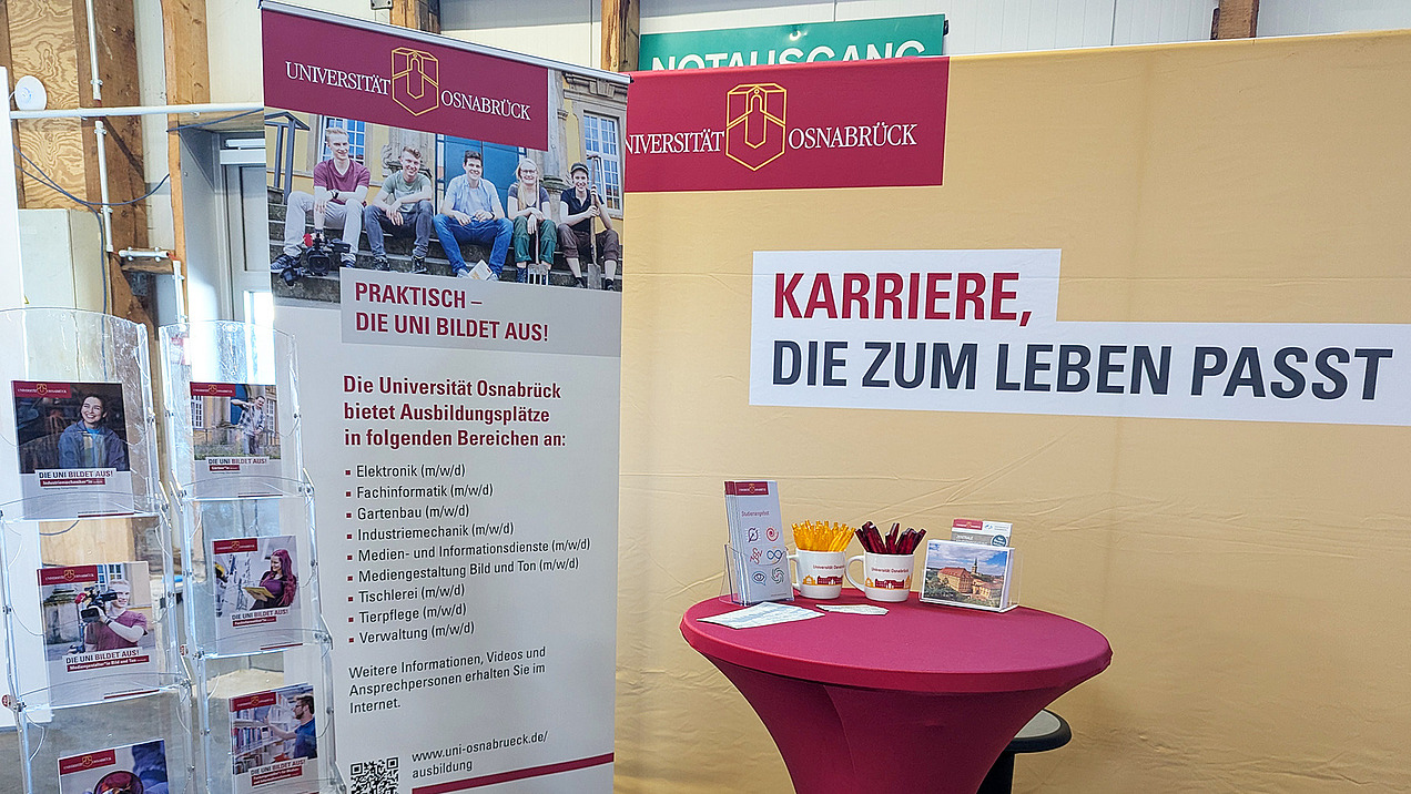 Stand der Universität Osnabrück auf einer Messe, der über Ausbildungsmöglichkeiten informiert. Im Fokus stehen Karrieremöglichkeiten, die zum Leben passen, und verschiedene Ausbildungsbereiche werden vorgestellt.