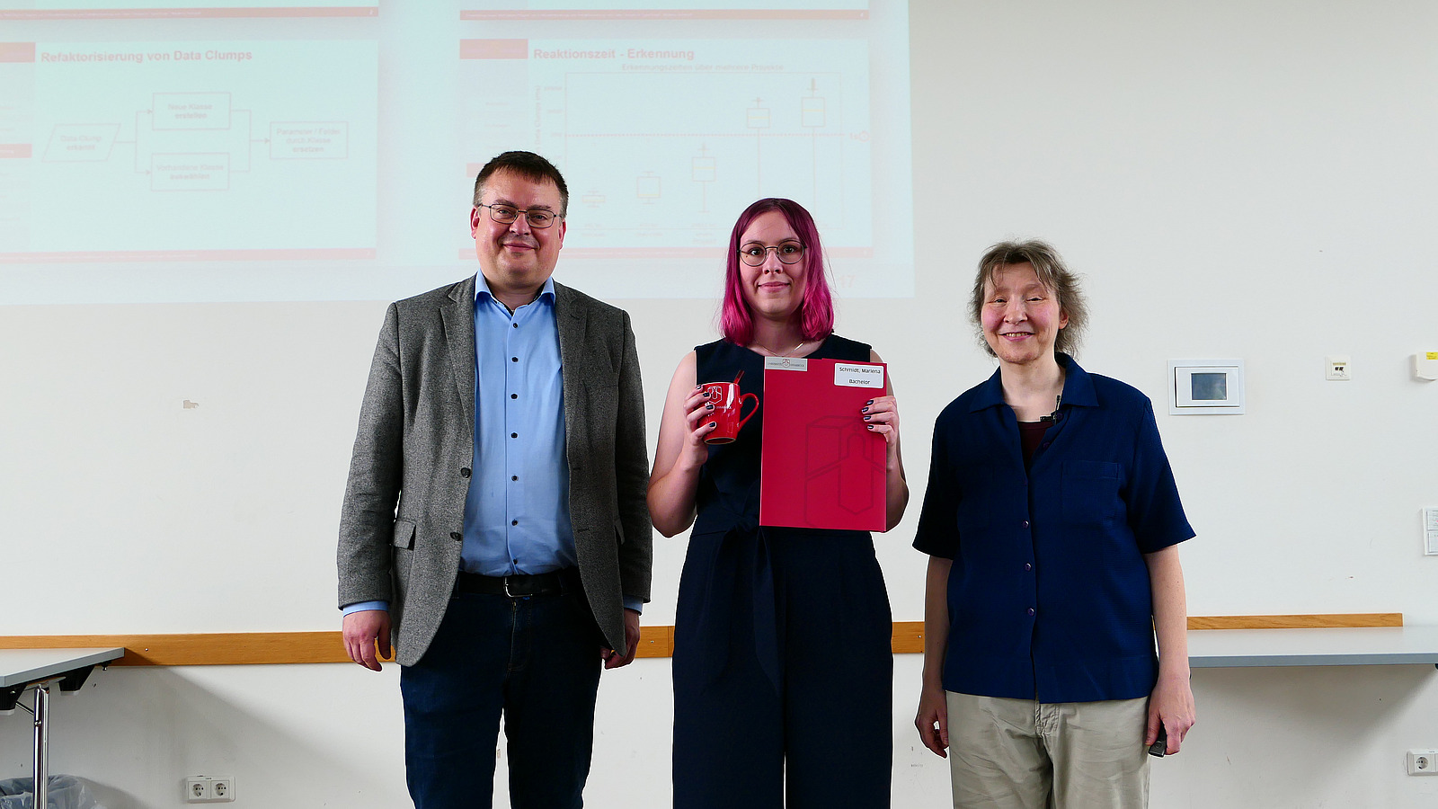 Eine Studentin mit Studiendekanin und Dekan stehen nebeneinander. Die Studentin hat eine Mappe in der Hand