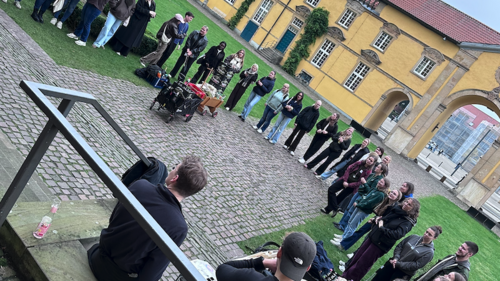 Mehrere Personen stehen in einem gro&szlig;en Kreis auf einem gepflasterten Innenhof. Einige sitzen am Rand auf dem Boden oder auf Stufen. In der Mitte des Kreises stehen Gegenst&auml;nde, darunter Taschen und weitere Materialien.  Im Hintergrund befindet sich ein gelbes Geb&auml;ude mit Fenstern und einer T&uuml;r. Neben dem gepflasterten Bereich ist eine Rasenfl&auml;che zu sehen. Im Vordergrund ist ein Gel&auml;nder sichtbar, dahinter sitzen zwei Personen.