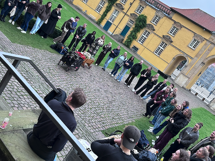 Mehrere Personen stehen in einem gro&szlig;en Kreis auf einem gepflasterten Innenhof. Einige sitzen am Rand auf dem Boden oder auf Stufen. In der Mitte des Kreises stehen Gegenst&auml;nde, darunter Taschen und weitere Materialien.  Im Hintergrund befindet sich ein gelbes Geb&auml;ude mit Fenstern und einer T&uuml;r. Neben dem gepflasterten Bereich ist eine Rasenfl&auml;che zu sehen. Im Vordergrund ist ein Gel&auml;nder sichtbar, dahinter sitzen zwei Personen.