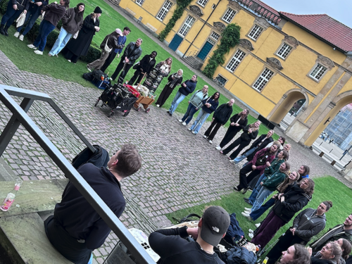 Mehrere Personen stehen in einem gro&szlig;en Kreis auf einem gepflasterten Innenhof. Einige sitzen am Rand auf dem Boden oder auf Stufen. In der Mitte des Kreises stehen Gegenst&auml;nde, darunter Taschen und weitere Materialien.  Im Hintergrund befindet sich ein gelbes Geb&auml;ude mit Fenstern und einer T&uuml;r. Neben dem gepflasterten Bereich ist eine Rasenfl&auml;che zu sehen. Im Vordergrund ist ein Gel&auml;nder sichtbar, dahinter sitzen zwei Personen.