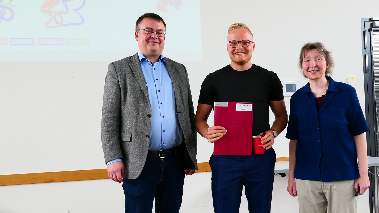 Ein Student mit Studiendekanin und Dekan stehen nebeneinander. Der Student hat eine Mappe in der Hand