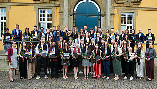 A group of people in festive attire stand in front of a historic building. They are all smiling at the camera. They are all wearing an elegant scarf and holding a flower and a certificate.