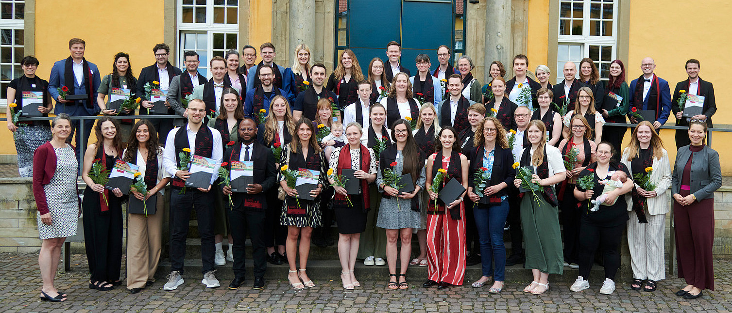 Eine Gruppe von Personen in festlicher Kleidung steht vor einem historischen Gebäude. Alle lächeln in die Kamera. Sie tragen alle einen eleganten Schal und halten eine Blume und eine Urkunde in der Hand.