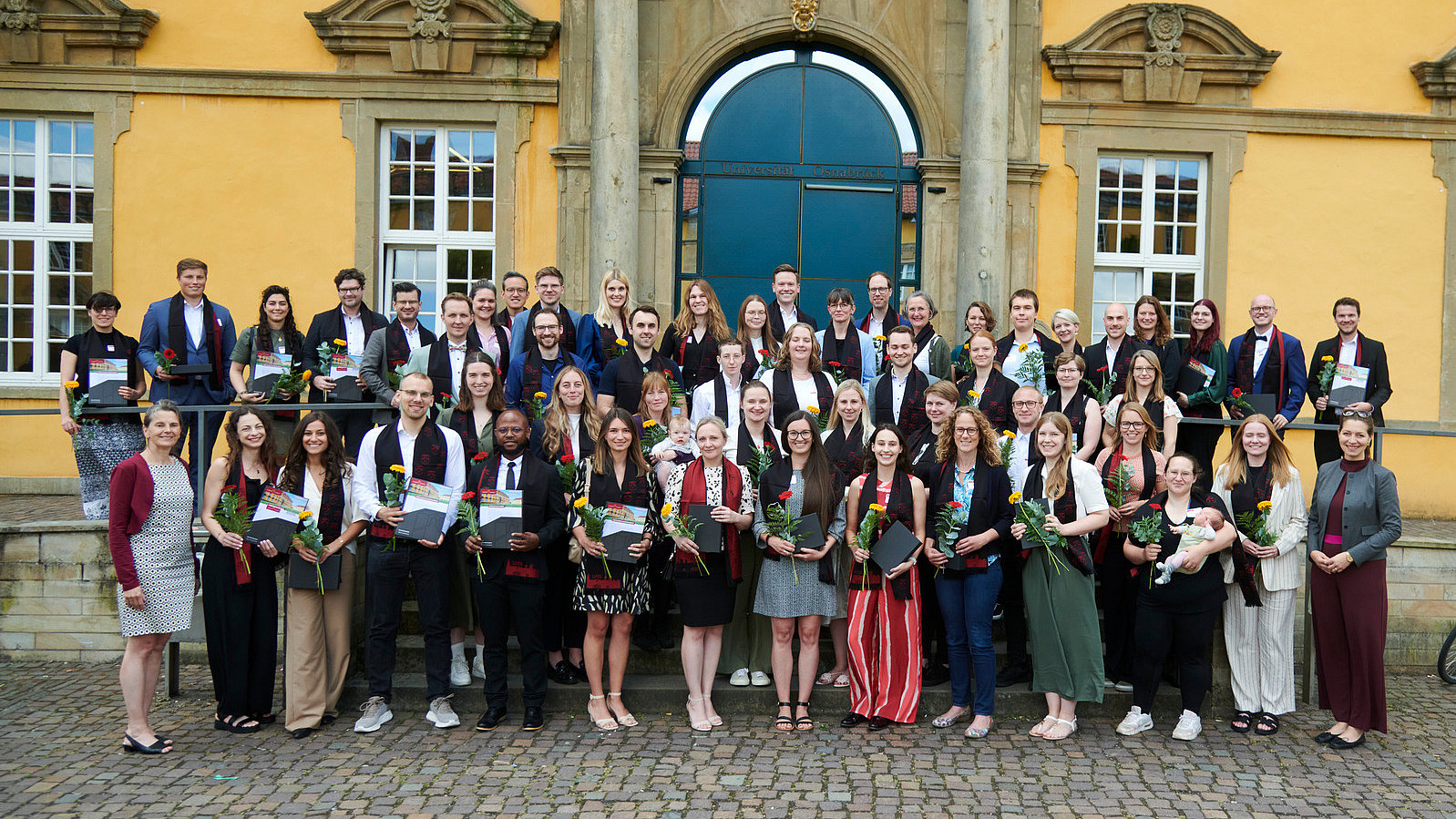 A group of people in festive attire stand in front of a historic building. They are all smiling at the camera. They are all wearing an elegant scarf and holding a flower and a certificate.