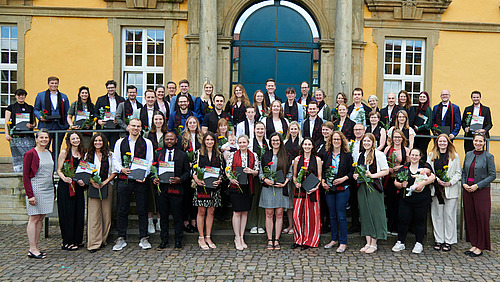 Eine Gruppe von Personen in festlicher Kleidung steht vor einem historischen Gebäude. Alle lächeln in die Kamera. Sie tragen alle einen eleganten Schal und halten eine Blume und eine Urkunde in der Hand.