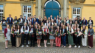 Eine Gruppe von Personen in festlicher Kleidung steht vor einem historischen Geb&auml;ude. Alle l&auml;cheln in die Kamera. Sie tragen alle einen eleganten Schal und halten eine Blume und eine Urkunde in der Hand.