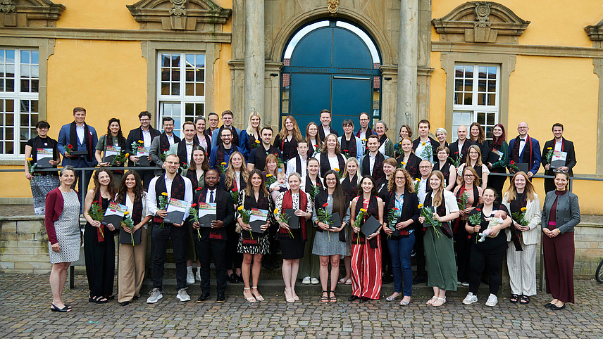 A group of people in festive attire stand in front of a historic building. They are all smiling at the camera. They are all wearing an elegant scarf and holding a flower and a certificate.