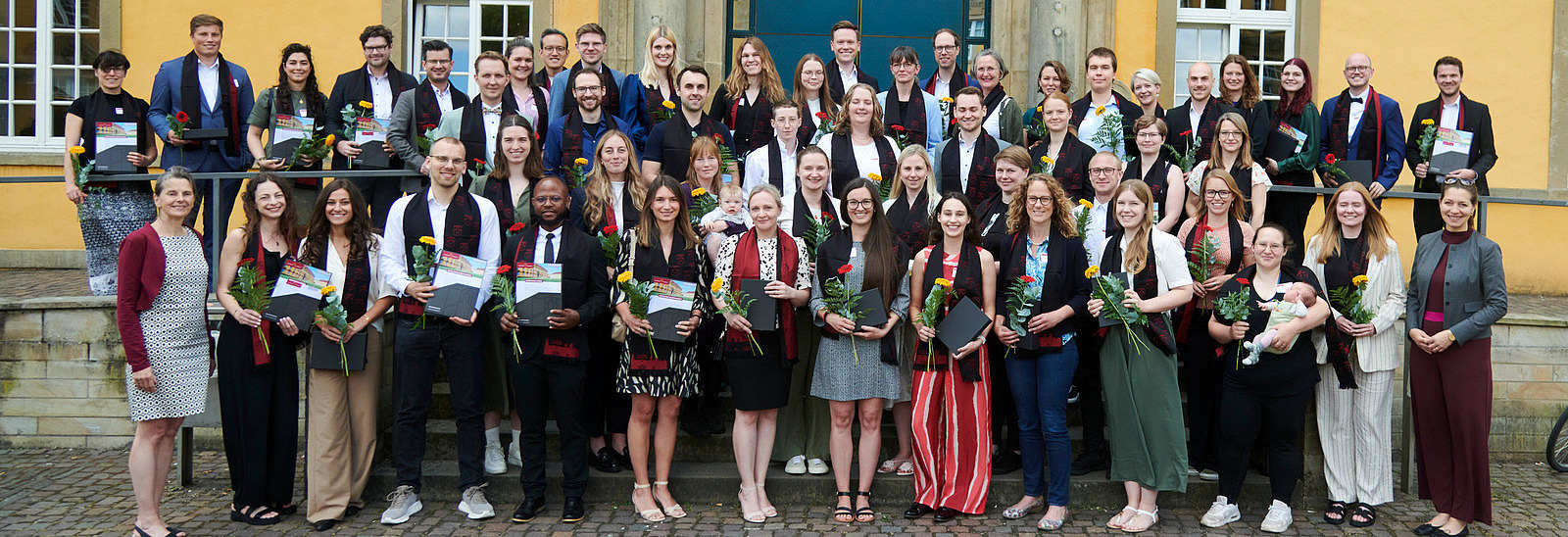 Eine Gruppe von Personen in festlicher Kleidung steht vor einem historischen Gebäude. Alle lächeln in die Kamera. Sie tragen alle einen eleganten Schal und halten eine Blume und eine Urkunde in der Hand.