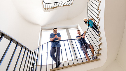 Drei Promoventen schauen in einem Treppenhaus auf den Betrachter herab.