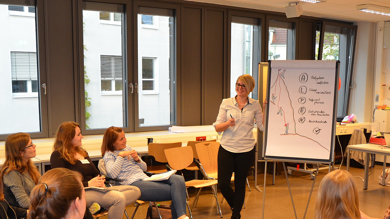 A lecturer stands at a flipchart. Students sit in a circle of chairs and listen.