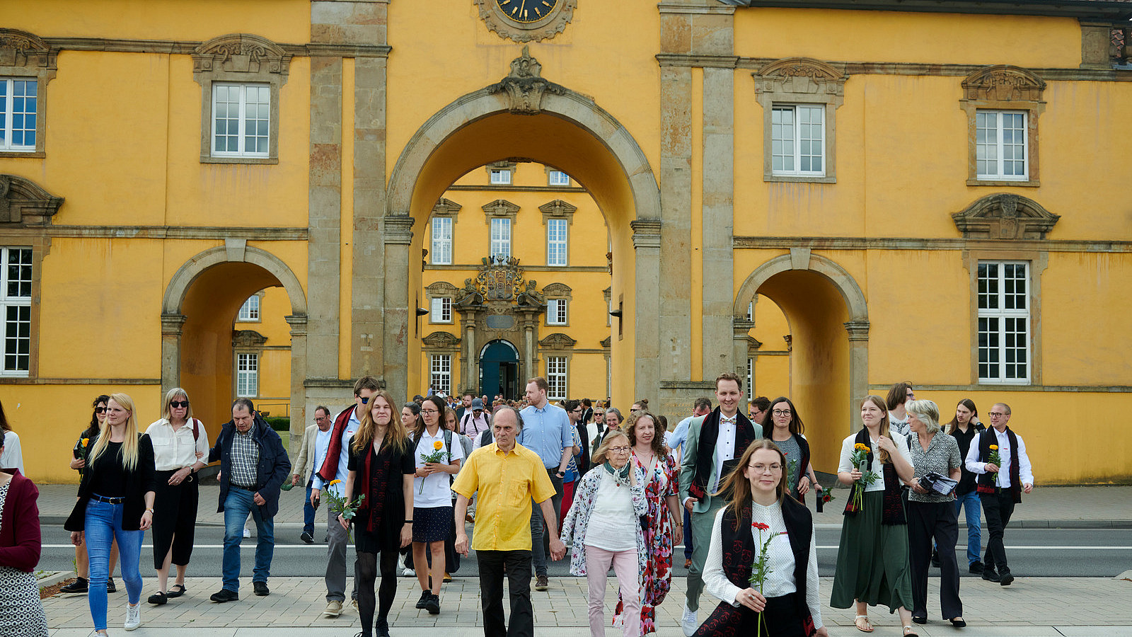 Numerous people enter the gate of a historic building, some cross the street in front of it