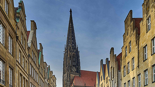 Eine Stra&szlig;e mit hohen, schmalen Geb&auml;uden auf beiden Seiten. Die Geb&auml;ude haben mehrere Stockwerke und unterschiedlich gestaltete Fassaden.  In der Mitte ragt ein hoher, spitzer Turm mit filigranen Strukturen in den Himmel. Hinter dem Turm ist ein rotes Dach zu sehen.  Der Himmel dar&uuml;ber ist bedeckt und grau.