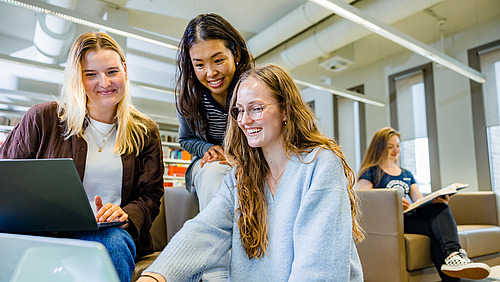 Drei junge Frauen in Bibliothek schauen gemeinsam auf einen Laptop.