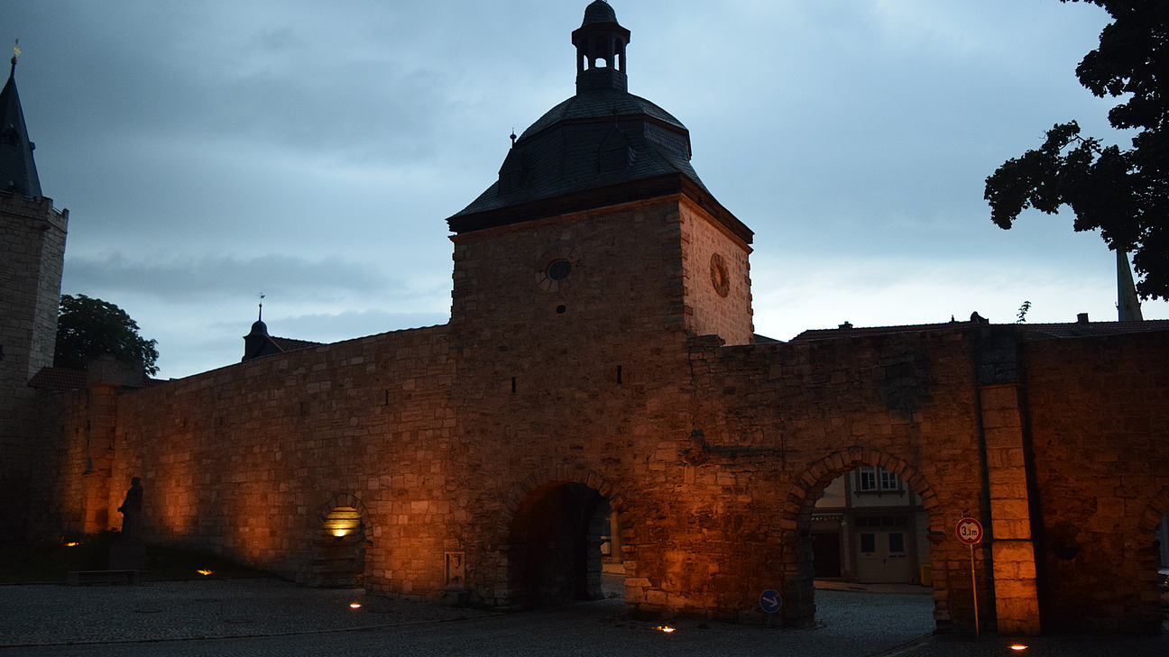 Das Bild zeigt das Tor einer mittelalterlichen Stadtmauer im Abendlicht. Das Tor ist in orangefarbenem Licht angestrahlt.