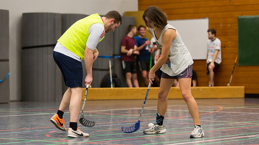 Ein Student und eine Studentin spielen zusammen Floorball in einer Sporthalle. Im Hintergrund stehen weitere Studierende.