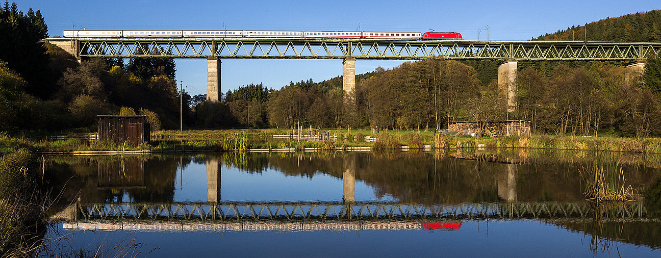 Fernzug InterCity fährt über eine Brücke und spiegelt sich mit blauem Himmel im Wasser wieder