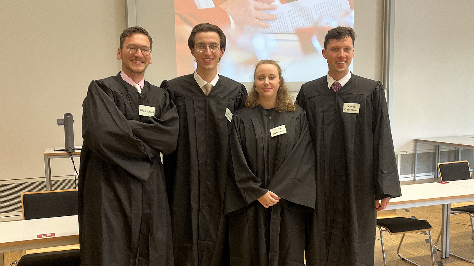 Eine Frau und drei M&auml;nner in schwarzen Roben mit grauen Namensschildern in einer Reihe stehend