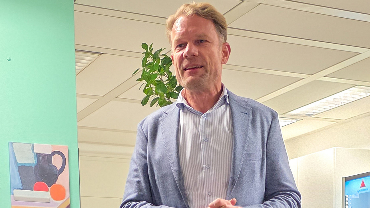 Ein Mann staht mit verschr&auml;nkten H&auml;nden in einem B&uuml;ro mit Pflanzen und einem Bild an der Wand